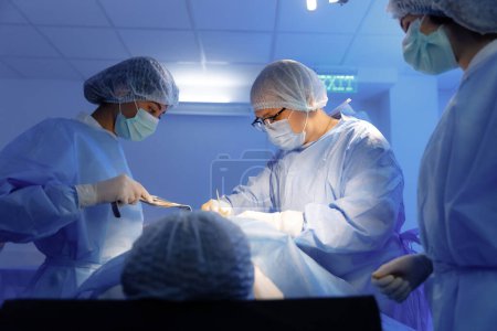 Téléchargez les photos : Deux infirmières et un chirurgien portant tous un gommage et du matériel chirurgical opèrent sur un patient et les infirmières surveillent très intensément pendant que le chirurgien effectue l'opération. Hôpital - en image libre de droit