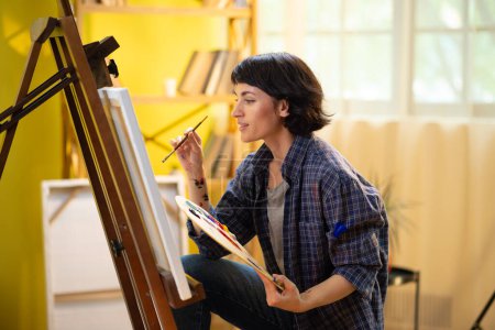 A very pretty woman with short brown hair is staring directly into the camera with her magnetizing eyes and sitting on a chair in her art apartment while painting something on a big canvas in front of