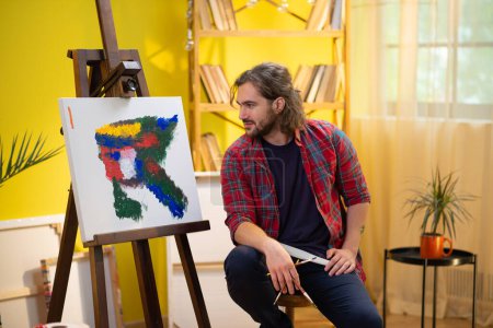 Foto de Un pintor guapo de pelo medio y barba está mirando su obra de arte y pintándola con su pincel y su paleta. - Imagen libre de derechos