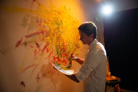 Téléchargez les photos : Bon artiste homme peignant une peinture moderne sur le mur de son atelier d'art il utilise la couleur à l'huile et le pinceau. Peinture - en image libre de droit