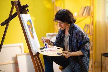 Foto de A beautiful lady with short hair is sitting down on a chair and painting something onto the canvas as she seems very relaxed. - Imagen libre de derechos