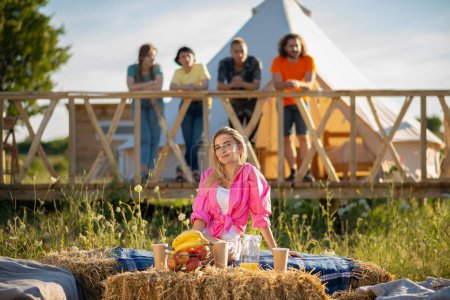 Foto de Camping moderno en un día de verano posando frente a la cámara hermosa dama mirando recto mientras sus amigos se enfrían en el fondo. - Imagen libre de derechos
