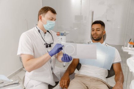Foto de Un odontólogo guapo con máscara protectora explica al paciente el proceso de trabajo que utiliza un modelo de dientes para mostrar y explicar más fácilmente. Dentista - Imagen libre de derechos
