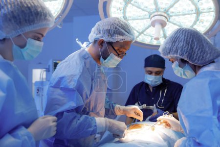 Photo for A room filled with medical equipment has three surgeons wearing blue scrubs and one other surgeon wearing a dark blue uniform discussing and trying to perform a surgery. Hospital - Royalty Free Image