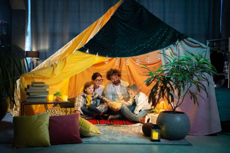 Téléchargez les photos : Devant la caméra grande famille deux enfants et les parents profiter de la nuit ensemble dans la tente faite à la main assis sur le sol dans la chambre des enfants manger du pop-corn et regarder un film sur l'ordinateur portable - en image libre de droit