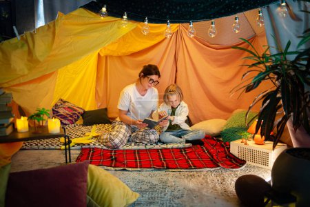 Photo for A higher camera shot of a modern and cozy looking indoor blanket tent where there is a wholesome moment of a mother and her son sharing a bonding time while reading together. Family - Royalty Free Image