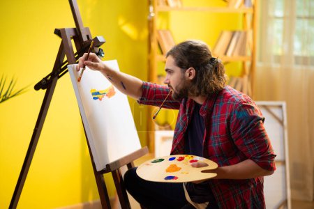 Foto de A painter handsome man is painting very carefully something beautiful on a big canvas in his art room. - Imagen libre de derechos