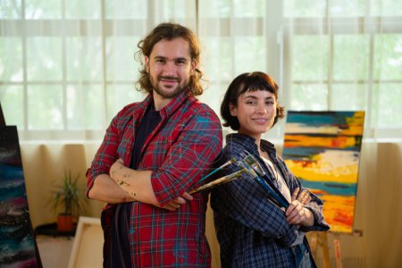 Téléchargez les photos : Attrayant et beau deux artiste posant devant la caméra dans le studio d'art ils tenant les pinceaux et croisant les mains en souriant mignon. Peinture - en image libre de droit