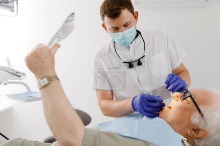 Téléchargez les photos : À la clinique dentaire le jeune dentiste regardant l'homme faisant un traitement pour les dents pour un vieil homme patient le patient tenant un miroir de main et regardant le processus. Dentiste - en image libre de droit