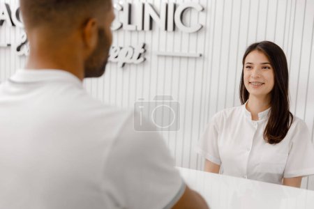 Téléchargez les photos : Jolie jeune assistante médicale à la réception discutant avec le client elle a invité le patient au cabinet du médecin elle sourit grand. - en image libre de droit