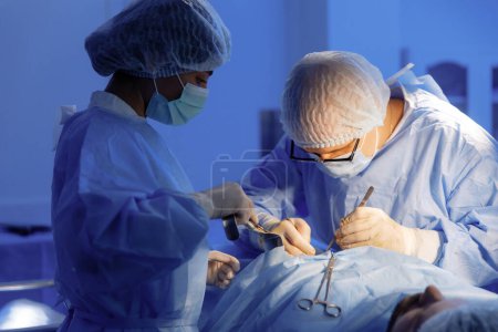 Foto de Un médico masculino con uniforme de cirujano completo está realizando una cirugía intensa mientras sus dos asistentes a cada lado de él lo están ayudando.. - Imagen libre de derechos