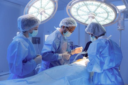 Téléchargez les photos : Quatre chirurgiens médicaux sont dans une pièce remplie de différents équipements médicaux travaillant sur un patient et se tenant autour de lui. - en image libre de droit