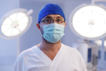Téléchargez les photos : Un médecin homme debout dans une salle d'opération portant des lunettes un filet à cheveux et un masque médical regardant dans la caméra alors qu'il se rapproche. - en image libre de droit