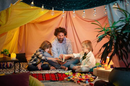 Photo for Two boys and their handsome father are wearing casual comfortable clothes and bonding together while reading a book and laughing in an indoor tent. - Royalty Free Image