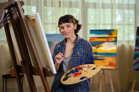 Photo for Creative young woman painter working on a new artwork for her project and posing in her art studio. Cheerful painter standing in front of her paintings. Paint - Royalty Free Image