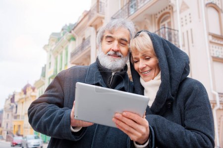 Téléchargez les photos : Attrayant mature couple caucasien debout dans la rue urbaine lecture d'informations sur un ordinateur tablette avec un sourire - en image libre de droit