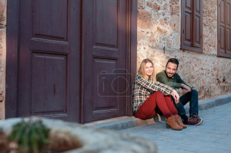 Photo for Young beautiful multiethnic couple sitting on the ground in city near old house and smiling - Royalty Free Image