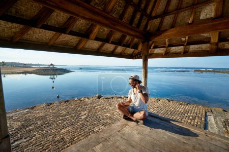 Photo for Relaxed and cheerful. Listening to music. Outdoor portrait of happy young african man resting on deck near the sea. - Royalty Free Image