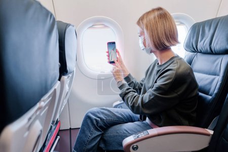 Téléchargez les photos : Une jeune femme portant un masque facial voyage en avion à l'aide d'un smartphone. Nouveau concept normal de voyage covid-19 pandémie. - en image libre de droit