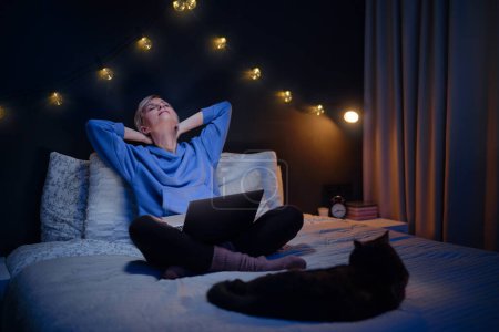 Foto de Mujer joven con su gato usando computadora portátil en la cama en su acogedor dormitorio por la noche. - Imagen libre de derechos