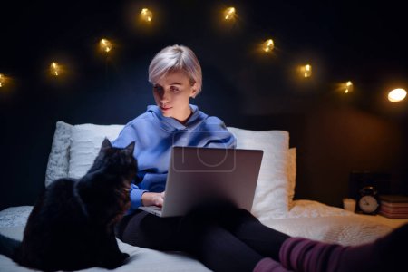 Photo for Young woman with her cat using laptop computer in bed in her cozy bedroom at night. - Royalty Free Image