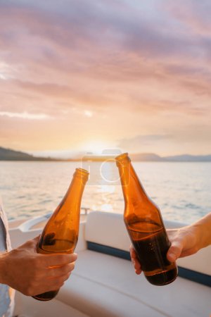 Foto de Fiesta con amigos. Primer plano de dos manos masculinas sosteniendo botellas de cerveza en el yate navegando por el mar. - Imagen libre de derechos