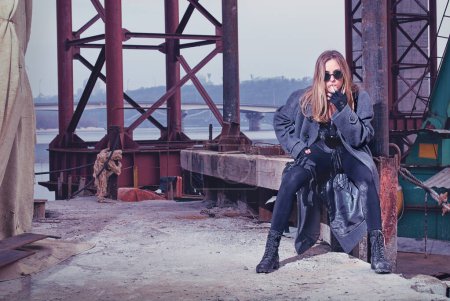 Foto de Chica armada en un gabardina enciende un cigarrillo mientras espera en un punto de encuentro bajo el puente cerca de un río. - Imagen libre de derechos
