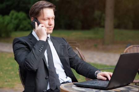 Téléchargez les photos : Un homme d'affaires concentré. attrayant jeune caucasien homme en formalwear travailler sur ordinateur portable tout en étant assis à la table à l'extérieur. - en image libre de droit