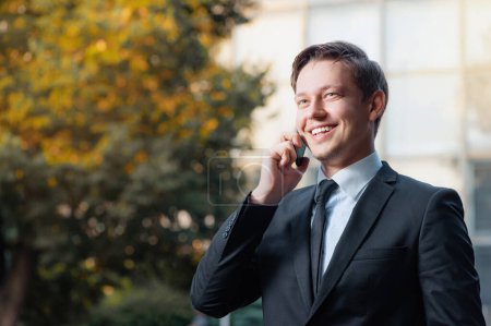 Téléchargez les photos : Entretien d'affaires confiant. Joyeux souriant caucasien jeune homme en tenue formelle parlant sur le téléphone mobile et regardant loin tout en se tenant debout à l'extérieur et contre la structure du bâtiment à l'extérieur - en image libre de droit