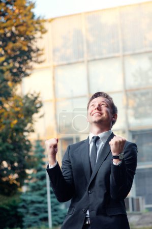 Téléchargez les photos : Gagnant. Heureux jeune homme caucasien en tenue de cérémonie gardant les bras levés et exprimant la positivité tout en se tenant debout à l'extérieur avec immeuble de bureaux en arrière-plan - en image libre de droit