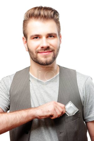 Téléchargez les photos : Soyez en sécurité ! Jeune homme souriant avec barbe mettant un préservatif dans sa poche de gilet. Isolé sur fond blanc. - en image libre de droit
