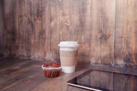 Foto de Trabajo en la cafetería. Tablet ordenador, taza de papel de café y magdalena en la mesa de madera. - Imagen libre de derechos