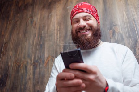 Funny news. Young laughing bearded man  holding smartphone against wooden wall.
