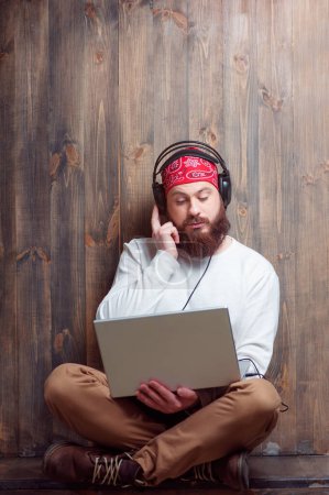 In the rhythm of my music. Relaxed young bearded man in headphones using laptop leaning on wooden wall and listening to the music.