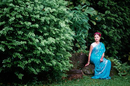 Foto de Hermosa mujer caucásica joven en ropa india tradicional con maquillaje nupcial y joyas. Bailarina de Bollywood en Sari y henna en las manos caminando por el jardín verde. - Imagen libre de derechos