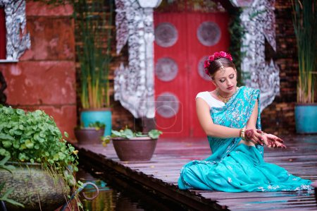 Téléchargez les photos : Belle jeune femme caucasienne en sari de vêtements indiens traditionnels avec maquillage nuptial et bijoux et tatouage au henné sur les mains dansant dans le jardin du temple
. - en image libre de droit
