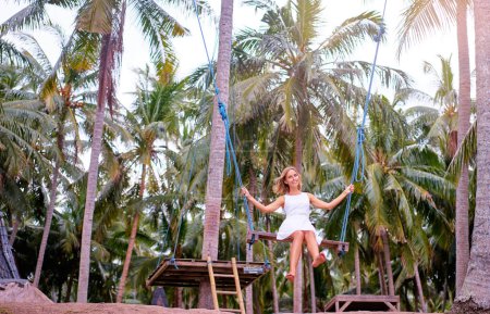 Photo for Vacation concept. Happy young woman in white dress swinging at palm grove. - Royalty Free Image