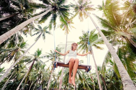 Foto de Concepto de vacaciones. Mujer joven feliz en vestido blanco y sombrero balanceándose en el palmeral
. - Imagen libre de derechos