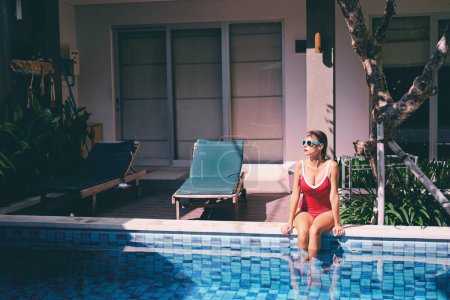 Photo for Enjoying suntan and vacation. Pretty young woman in red swimsuit sitting near swimming pool. - Royalty Free Image