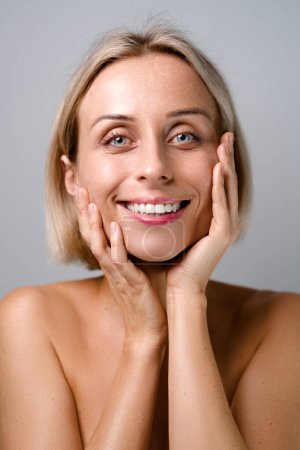 Skin care. Blonde woman with beauty face touching healthy facial skin. No makeup. Grey background.