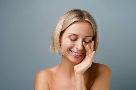 Skin care. Blonde woman with beauty face touching healthy facial skin. No makeup. Grey background.