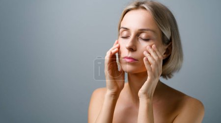 Cuidado de la piel. Mujer rubia con la cara de belleza tocando la piel facial saludable. Sin maquillaje. Fondo gris.