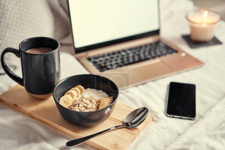 Photo for Breakfast in bed: a wooden tray with granola and cup of cocoa with a laptop on background. - Royalty Free Image