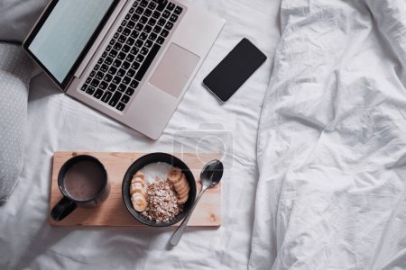 Foto de Desayuno en la cama: una bandeja de madera con granola y una taza de cacao con un ordenador portátil. - Imagen libre de derechos