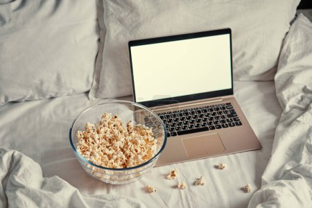 Téléchargez les photos : Regarder un film à la maison. Popcorn dans le bol et ordinateur portable au lit. Concept de divertissement. - en image libre de droit
