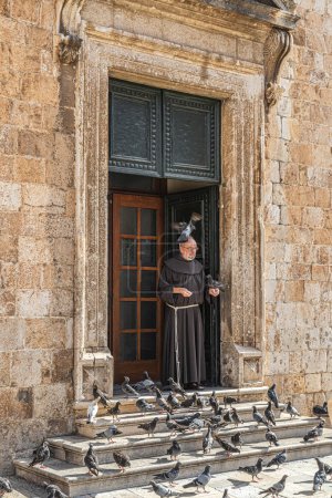 Photo for DUBROVNIK, CROATIA-AUGUST 18, 2022: Funny situations with a monk very friendly feeding the pigeons in front of the Franciscan Monastery and Church (Croatian: Franjevacki samostan i crkva u Dubrovniku) - Royalty Free Image