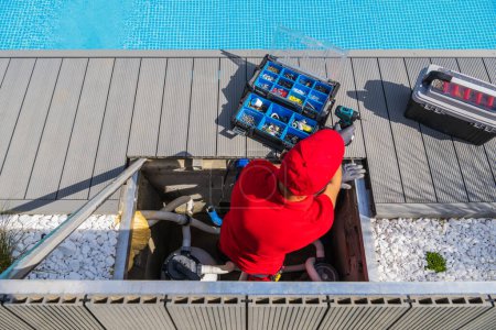 Professioneller Techniker in roter Uniform, der regelmäßige Wartungsarbeiten am Pool durchführt. Öffnen Sie die Werkzeugkiste mit verschiedenen Werkzeugen und Komponenten vor ihm. Ansicht von oben.