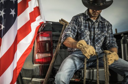 Cowboy Rancher caucasien dans la quarantaine assis sur son lit de camionnette avec une corde dans les mains. Préparation d'outils pour certains travaux de ranch. Drapeau américain à côté de lui.