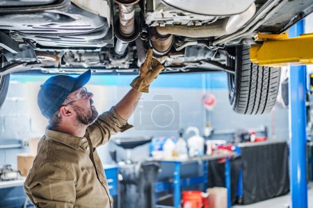 Professional Caucasian Mechanic Standing Under the Vehicle Lifted on Car Lift Checking the Condition of Catalytic Converter. Automotive Theme.