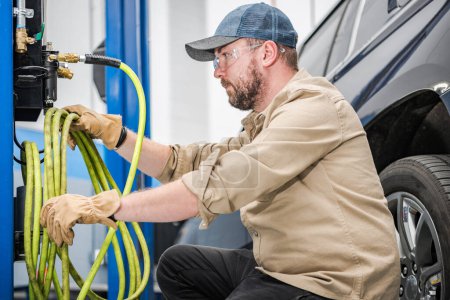 Foto de Caucasian Middle Aged Mechanic Taking Compressor Cables to Inflate Vehicle Tires After Seasonal Change. Professional Car Maintenance Station. - Imagen libre de derechos